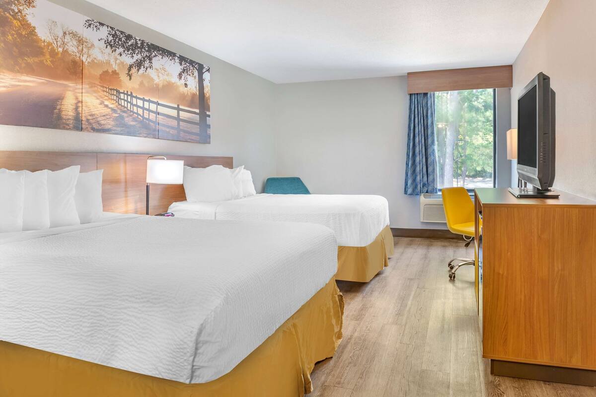 Ein Hotelzimmer mit zwei Betten, einem Schreibtisch, einem Fernseher und einem großen Fenster mit Vorhängen.