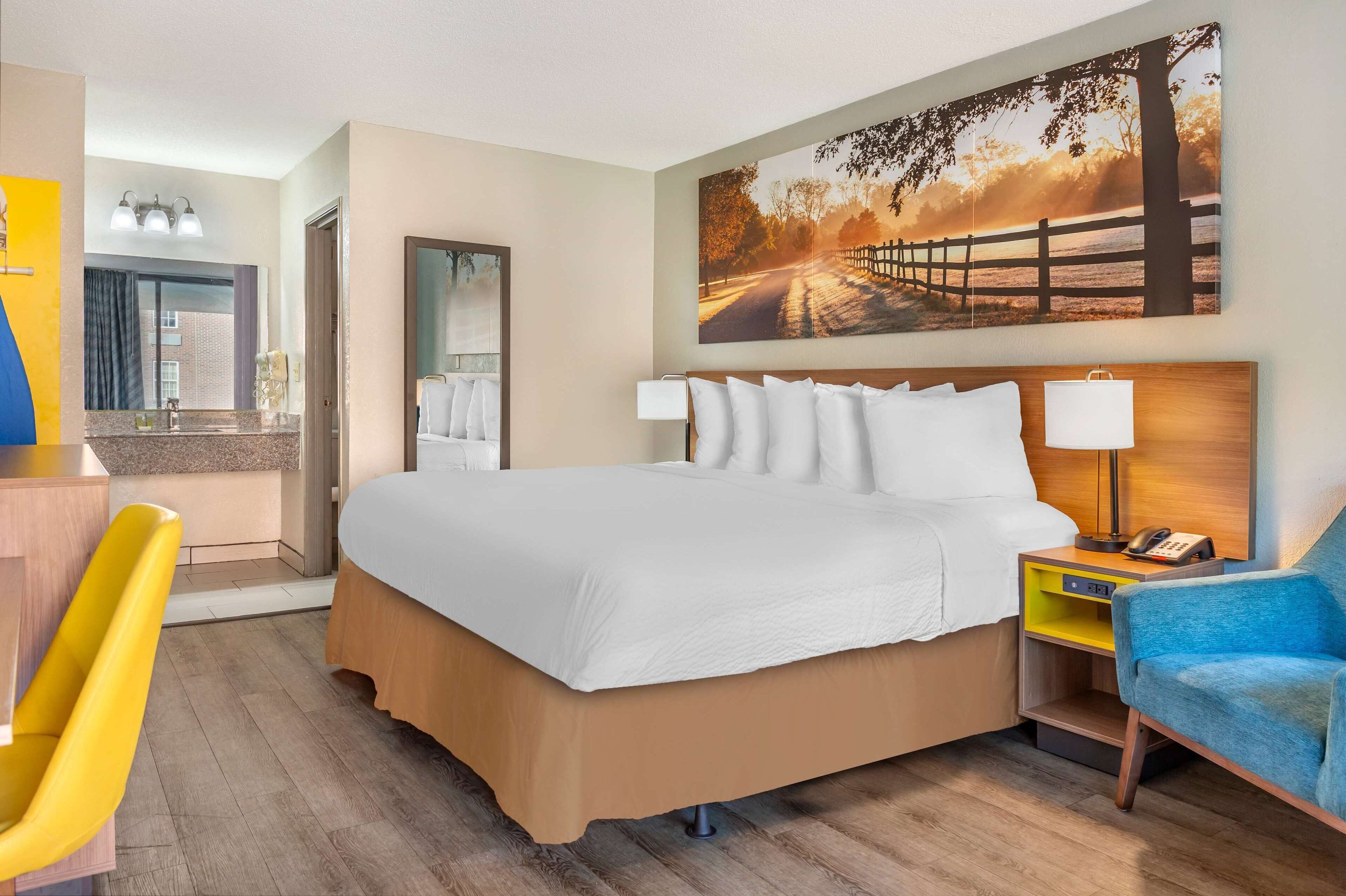 Een hotelkamer met een groot bed, een bureau met stoel, een spiegel, een schilderij van een zonsondergang en een nachtkastje met lamp.