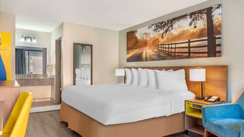 Een hotelkamer met een groot bed, een bureau met stoel, een spiegel, een schilderij van een zonsondergang en een nachtkastje met lamp.