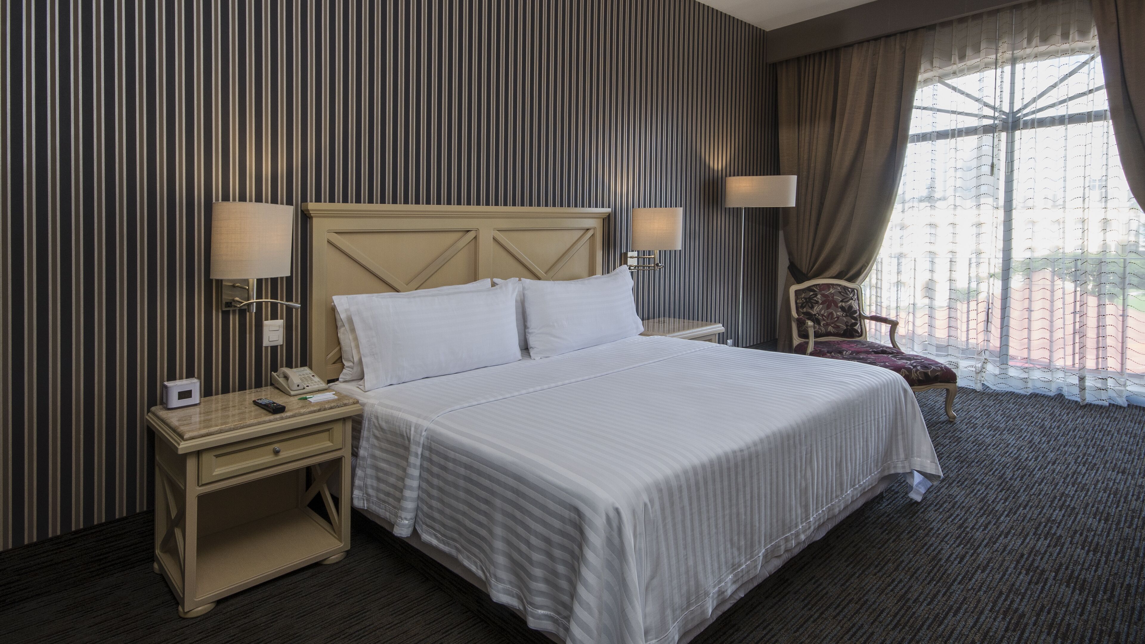Une chambre d’hôtel avec un grand lit, une table de chevet avec un téléphone et une lampe, une chaise et une fenêtre avec des rideaux.