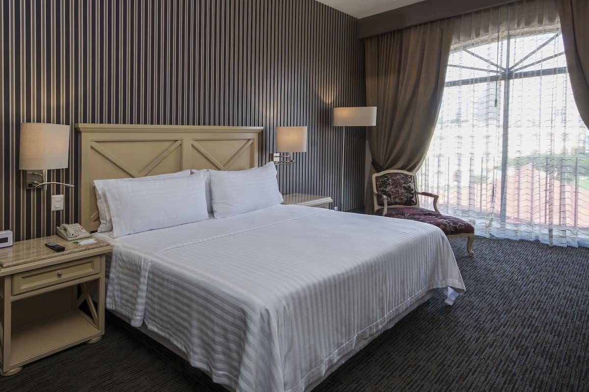 Habitación de hotel con una cama grande, una mesita de noche con teléfono y lámpara, una silla y una ventana con cortinas.