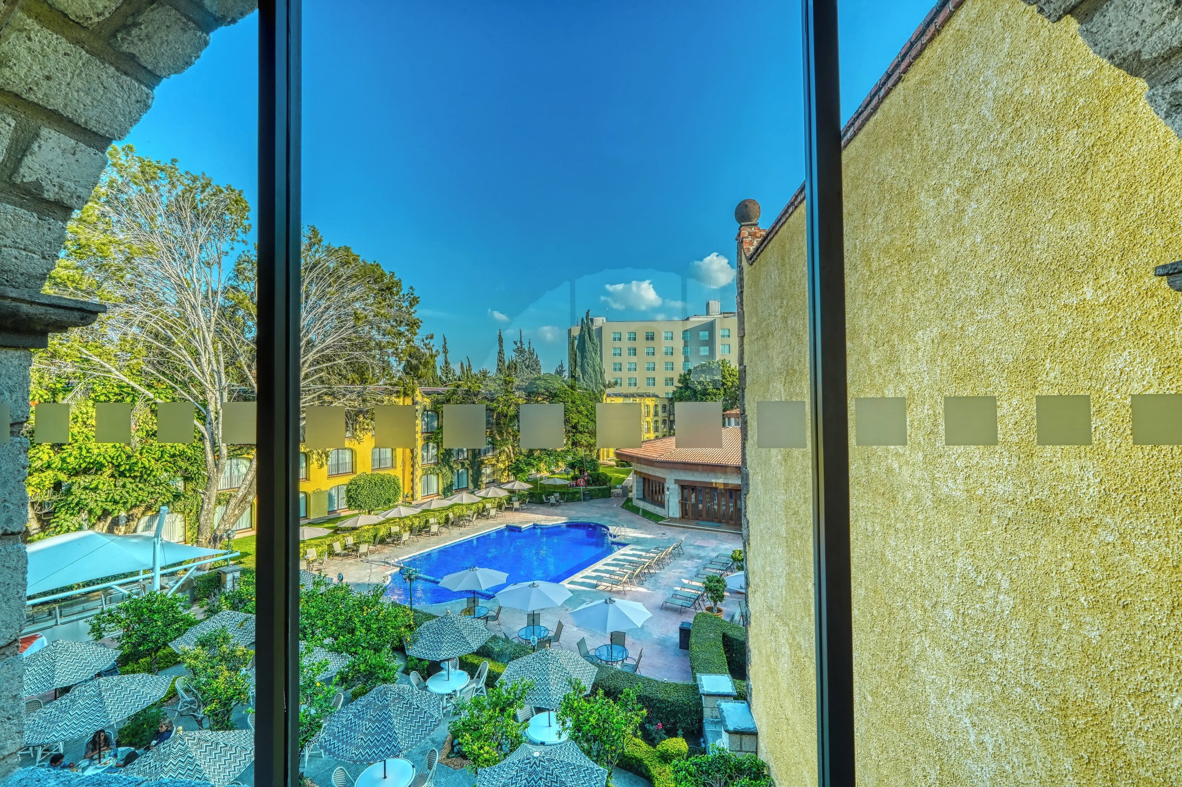 Zona de piscina con sillones de descanso y sombrillas, rodeada de follaje y edificios.
