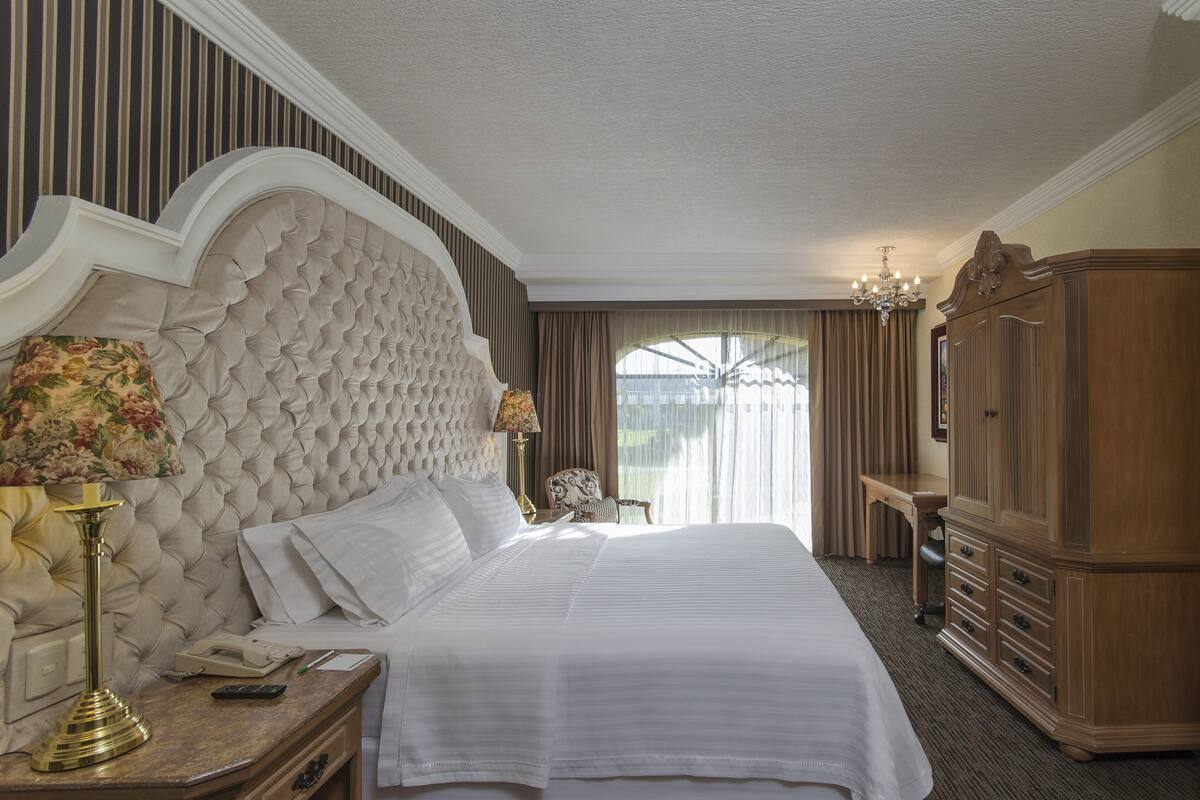 A hotel room with a large bed, a wooden wardrobe, a desk with a lamp, and a painting on the wall.
