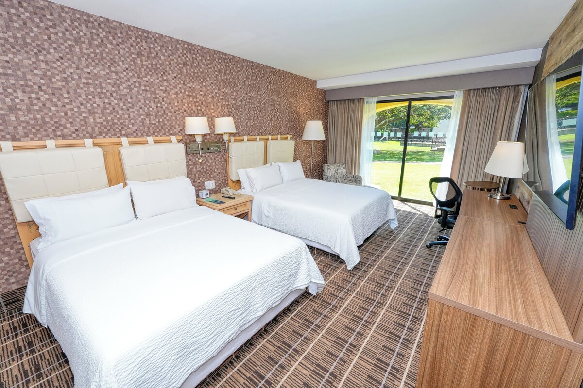A hotel room with two beds, a desk, a chair, and a view of the outdoors.
