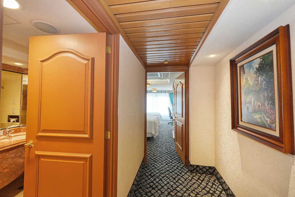 A hotel hallway with a wooden door, a patterned carpet, a bed, and a framed picture on the wall.