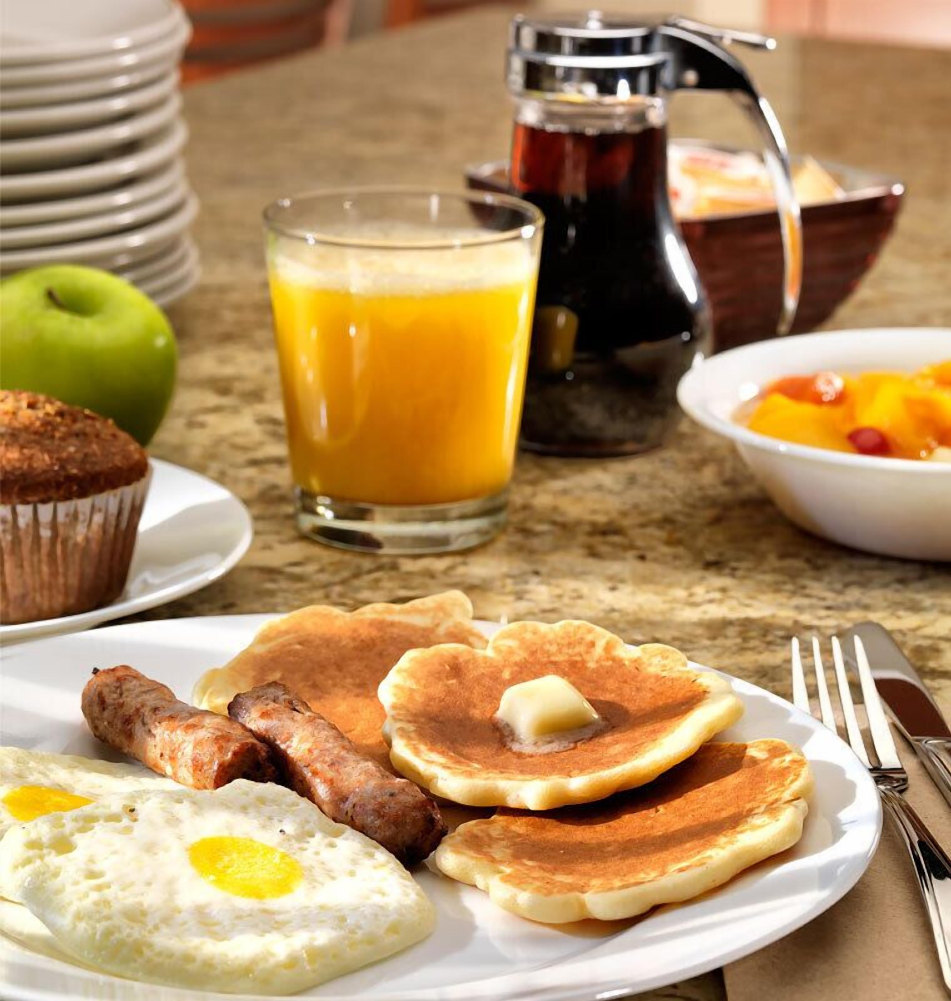 A breakfast plate with pancakes, sausages, and eggs, a glass of orange juice, a muffin, and a bowl of fruit.