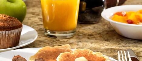 A breakfast plate with pancakes, sausages, and eggs, a glass of orange juice, a muffin, and a bowl of fruit.
