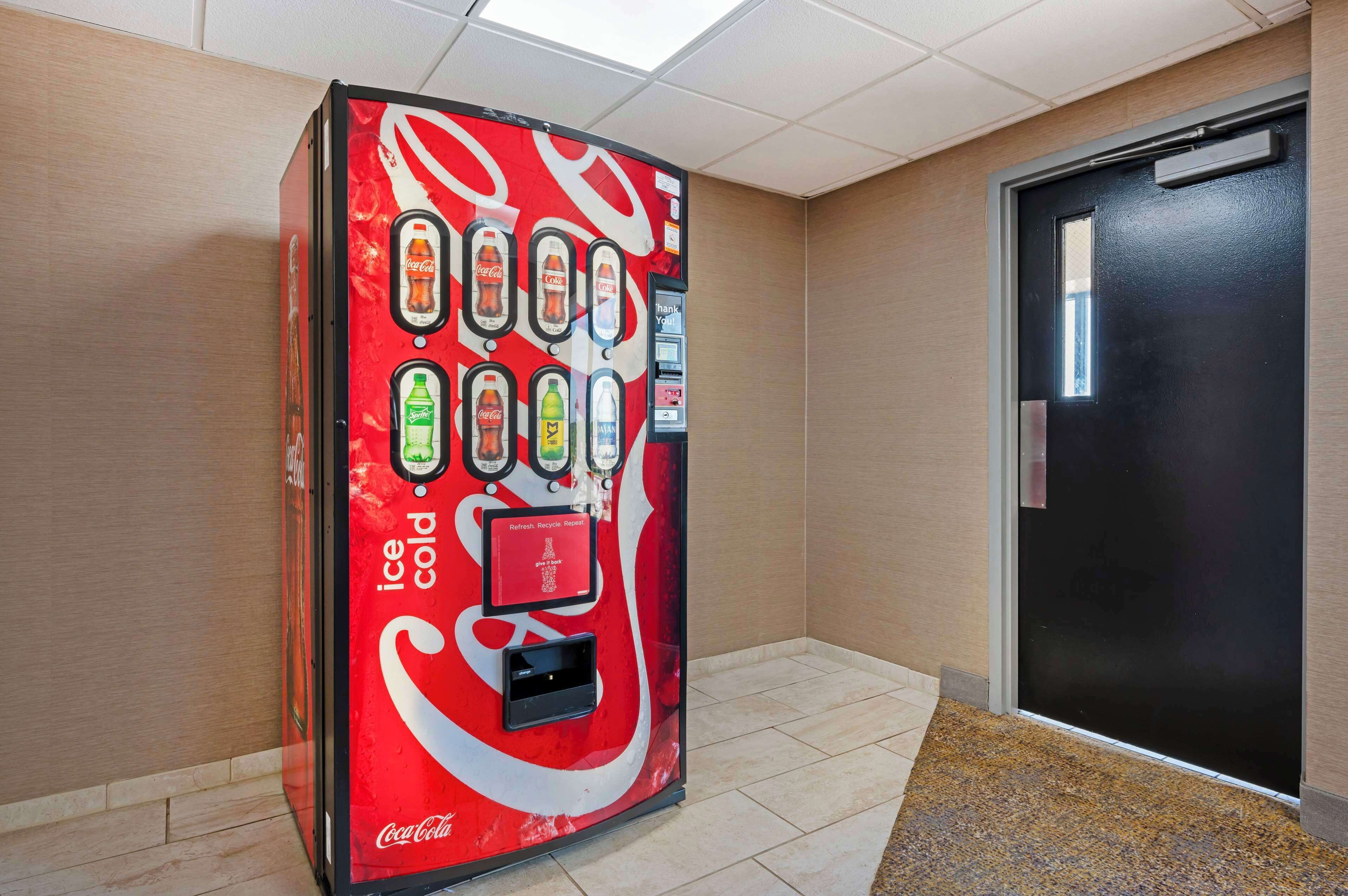 Una máquina expendedora de Coca-Cola en un pasillo con suelo de baldosas y una puerta al fondo.