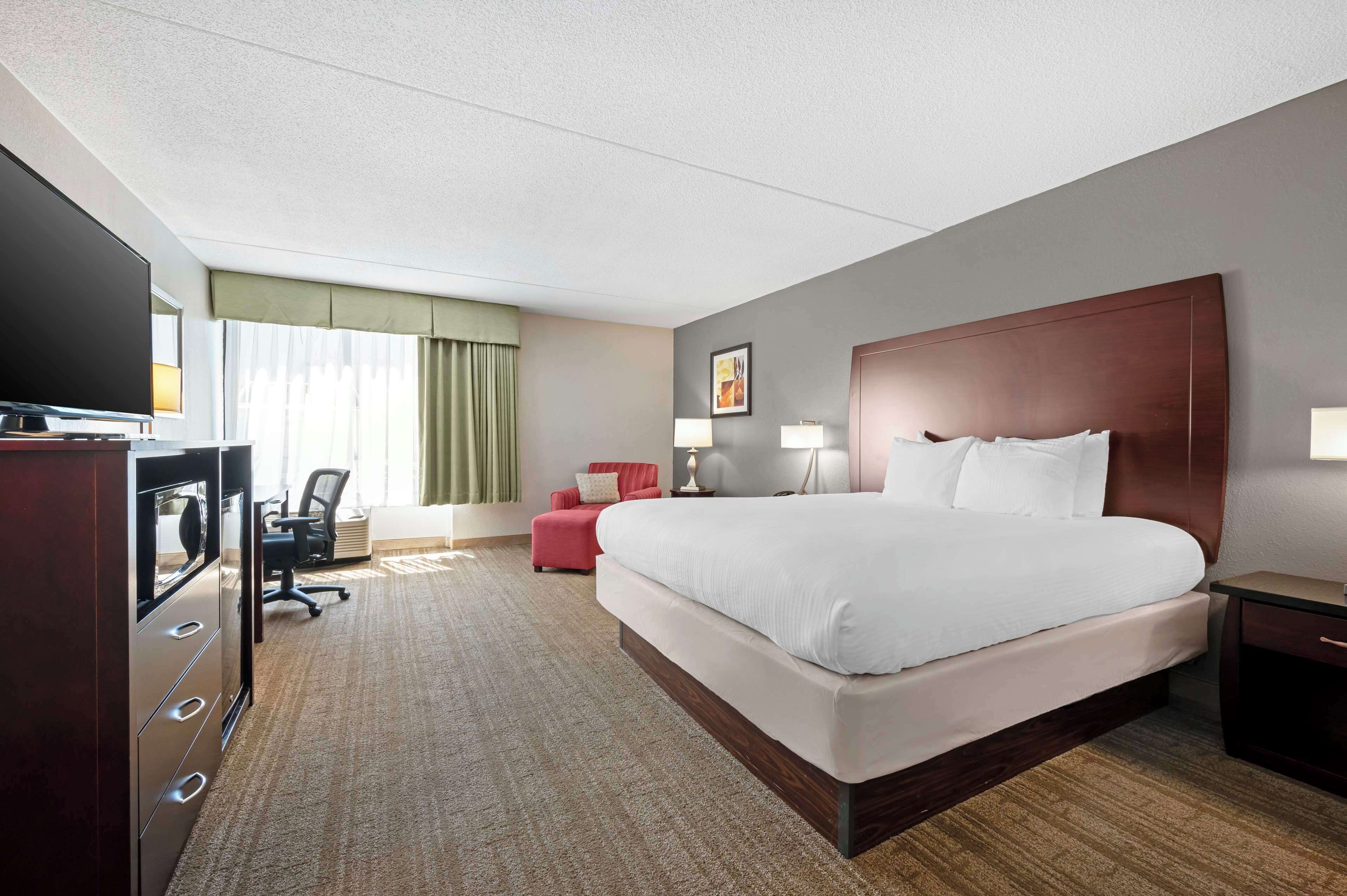 A hotel room with a large bed, a desk with a flat-screen TV, a red armchair, and a lamp.