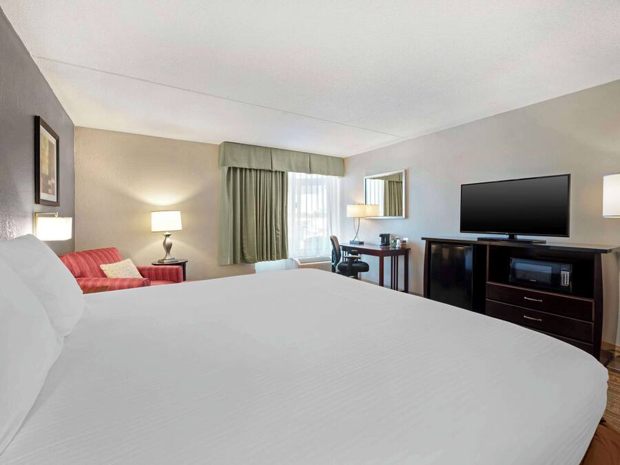 A hotel room with a large bed, a desk with a flat-screen TV, a chair, a lamp, a red sofa, and a window with curtains.