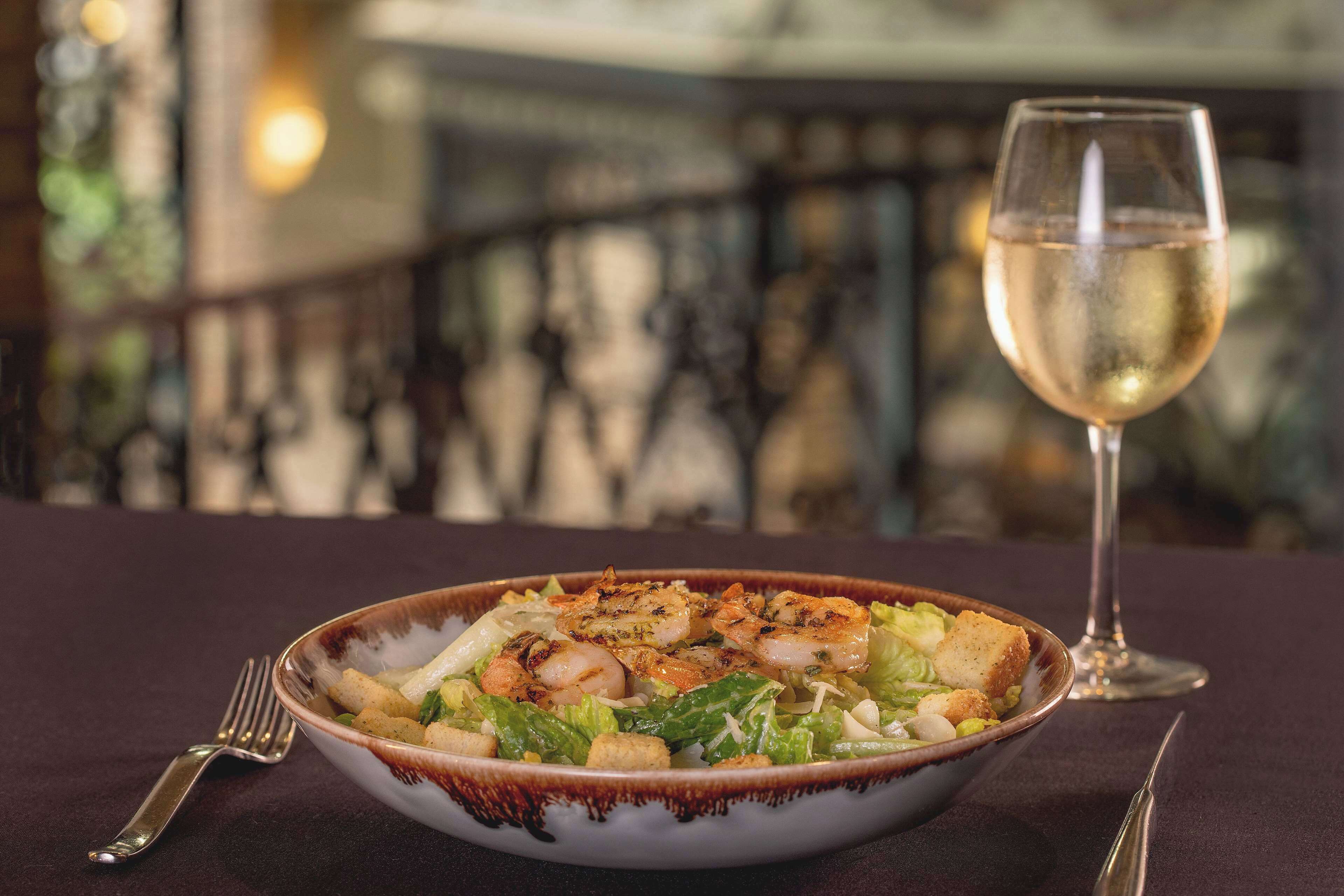 A bowl of salad with croutons and a glass of white wine on a table.