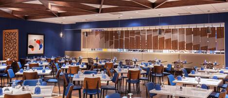 Un vaste espace dédié à la salle à manger, avec de nombreuses tables et chaises, un plafond haut orné de poutres en bois et un grand mur décoré d’un motif géométrique.