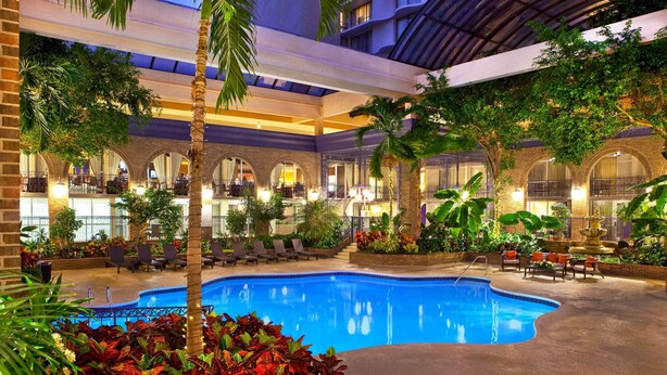 An indoor swimming pool with lounge chairs and palm trees, surrounded by a multi-level building with arched windows and a covered terrace. - Courtland Grand Hotel, Trademark Collection by Wyndham (Atlanta)