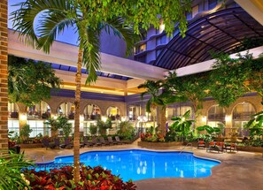 An indoor swimming pool with lounge chairs and palm trees, surrounded by a multi-level building with arched windows and a covered terrace. - Courtland Grand Hotel, Trademark Collection by Wyndham (Atlanta)