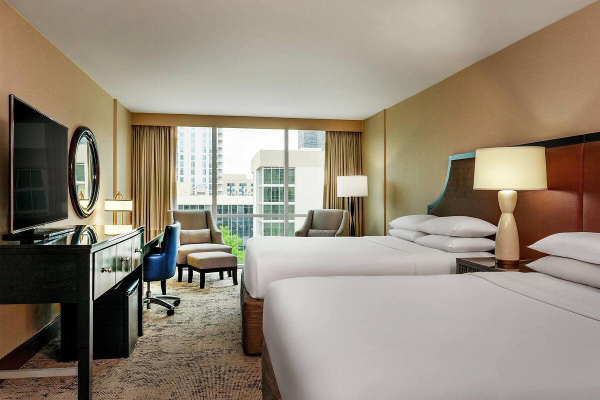 A hotel room with two beds, a desk with a TV, a chair, a lamp, and a view of the city.