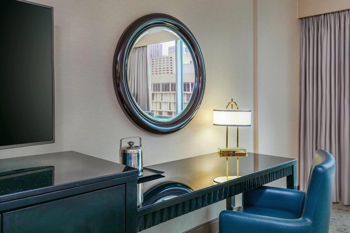 A hotel room with a large mirror, a desk with a lamp, a chair, and a view of buildings through the window.