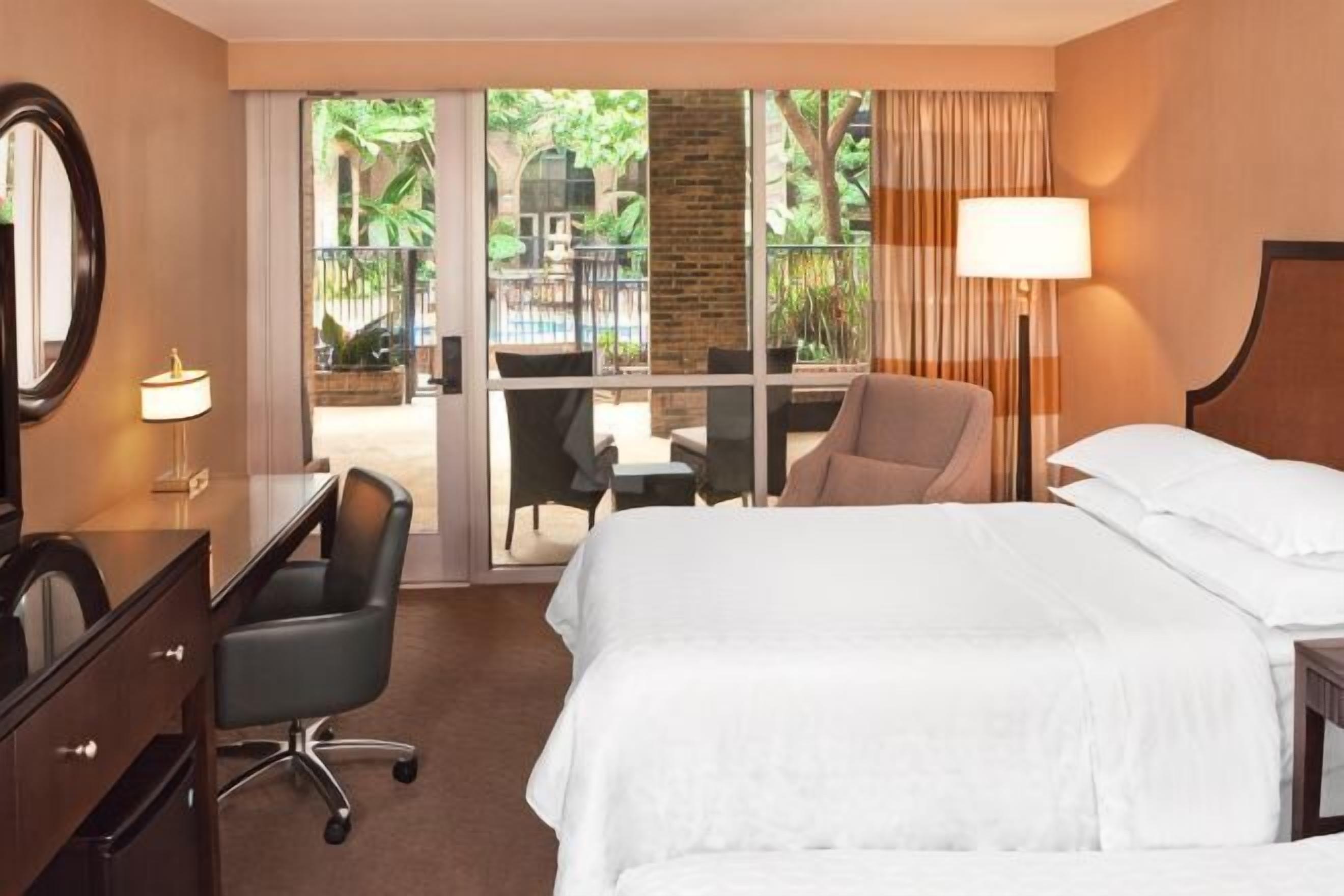 Une chambre d’hôtel équipée d’un lit, d’un bureau, d’une chaise et offrant une vue sur un espace extérieur avec une piscine et de la verdure.