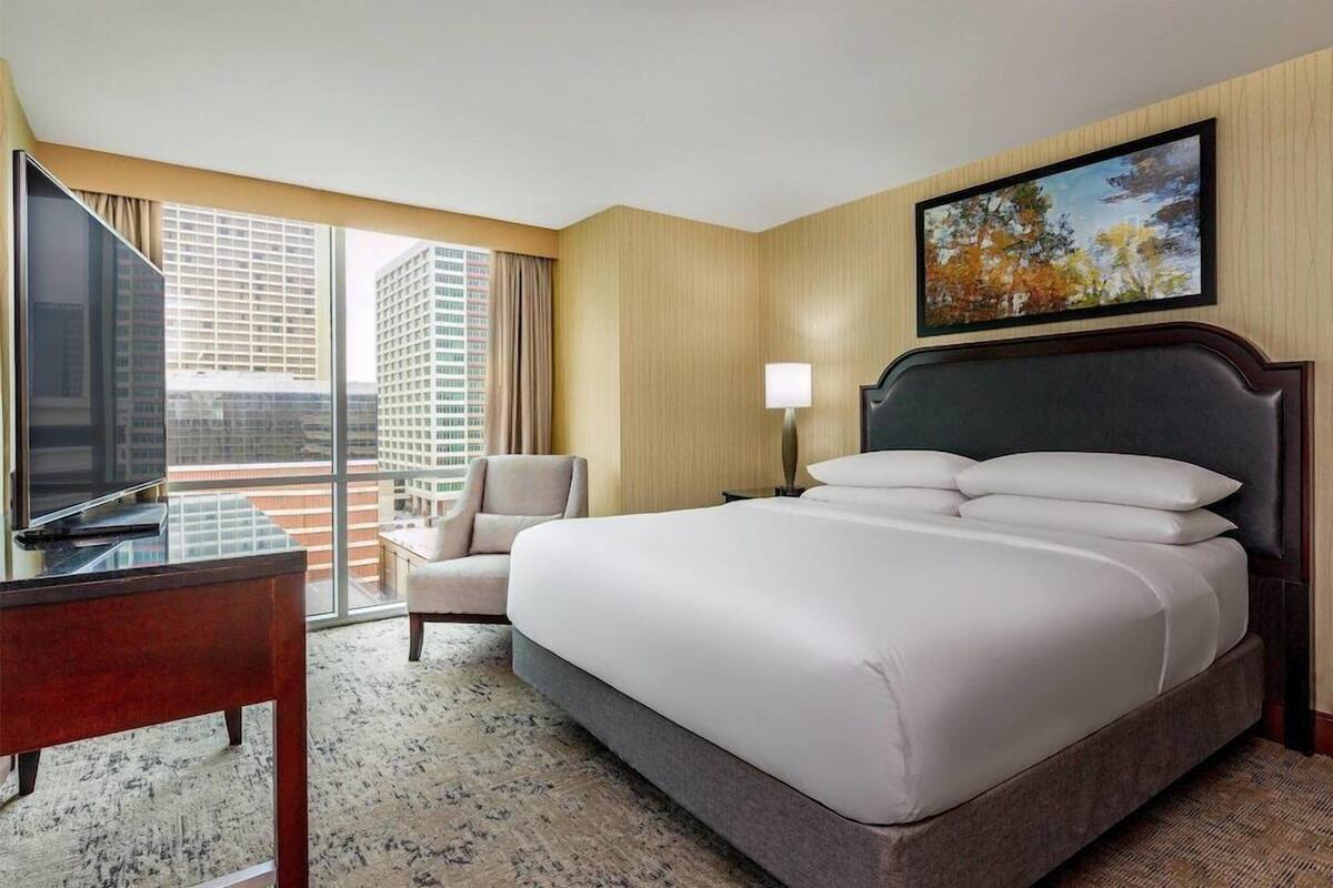 A hotel room with a large bed, a desk, a chair, a TV, and a window with a city view.