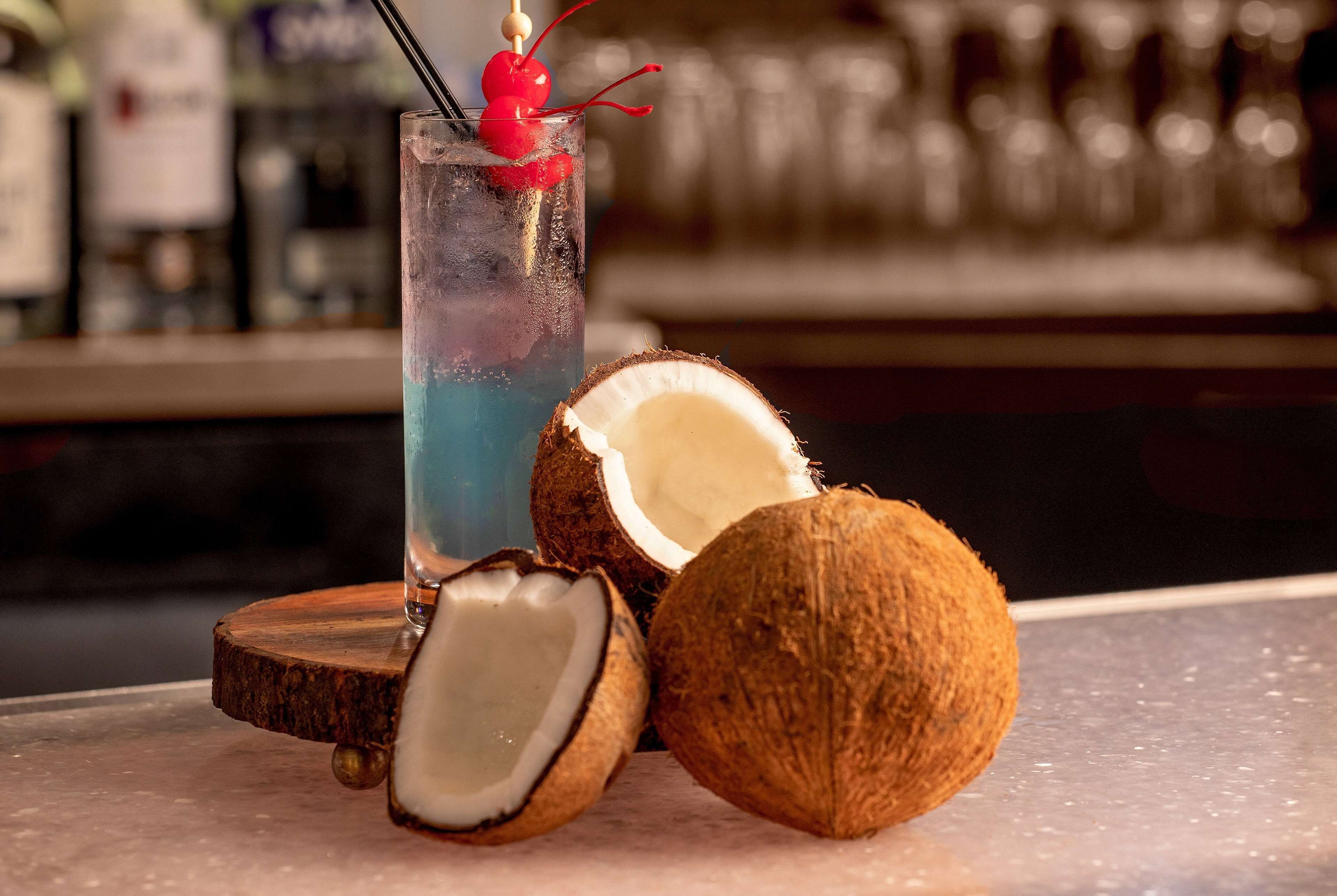 A cocktail glass with a blue liquid and a cherry on a stick, a half-cut coconut, and a wooden coaster.