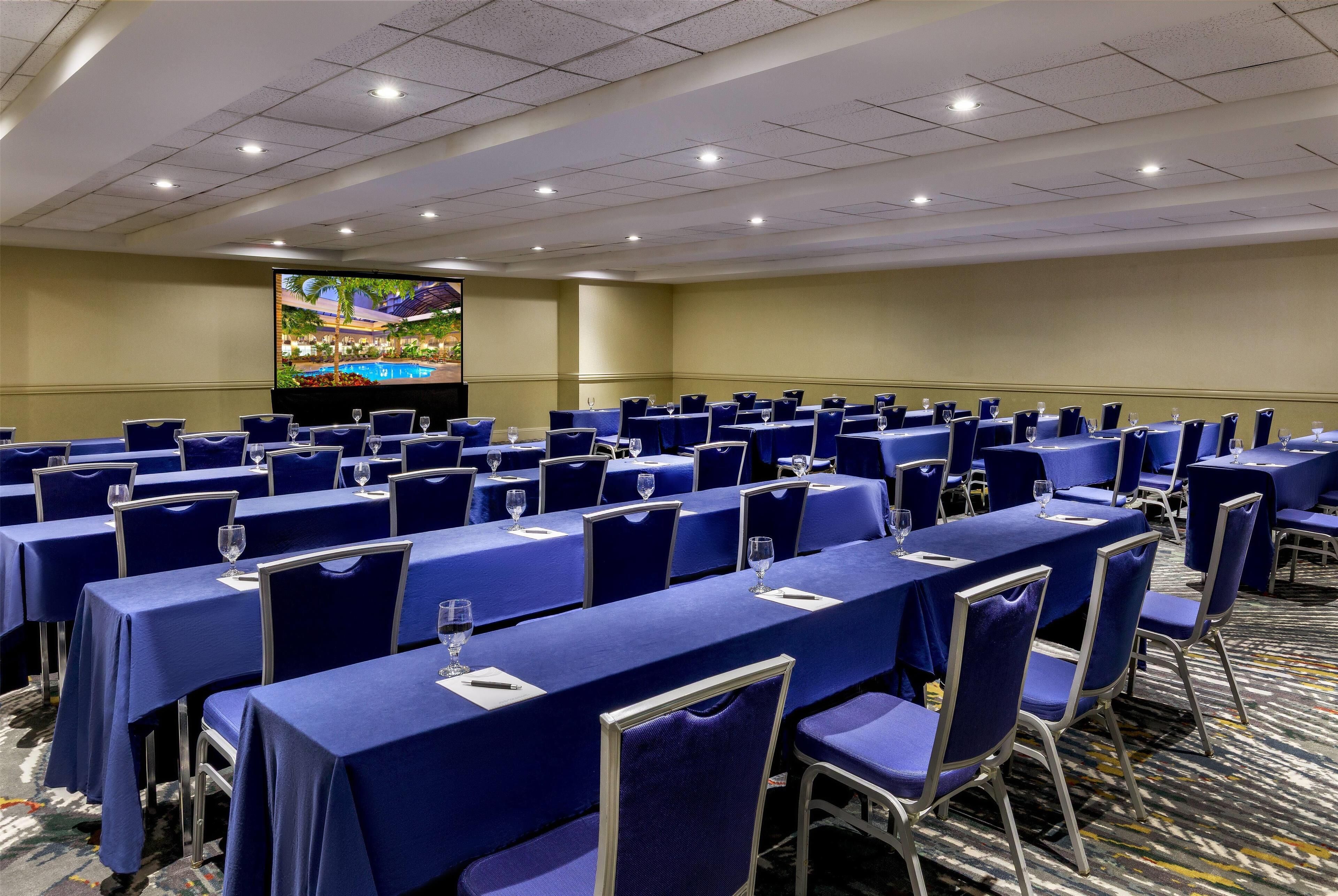 A conference room with rows of tables and chairs, a large screen, and a carpeted floor.