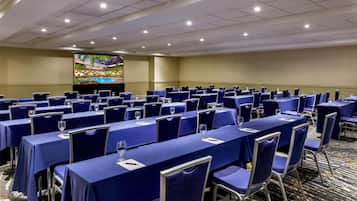A conference room with rows of tables and chairs, a large screen, and a carpeted floor.