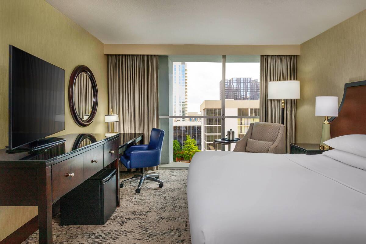 A hotel room with a bed, a desk, a chair, a TV, and a view of the city through large windows.