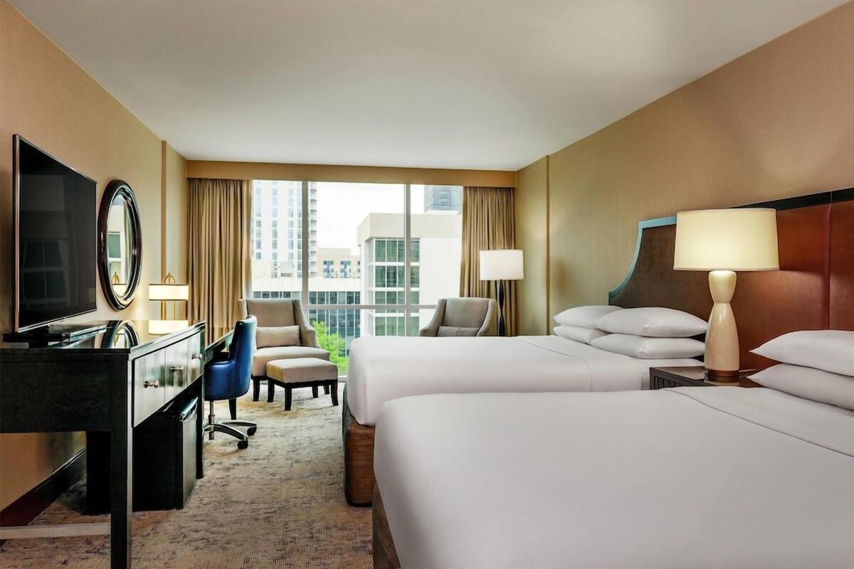 A hotel room with two beds, a desk, a chair, a TV, and a view of the city.