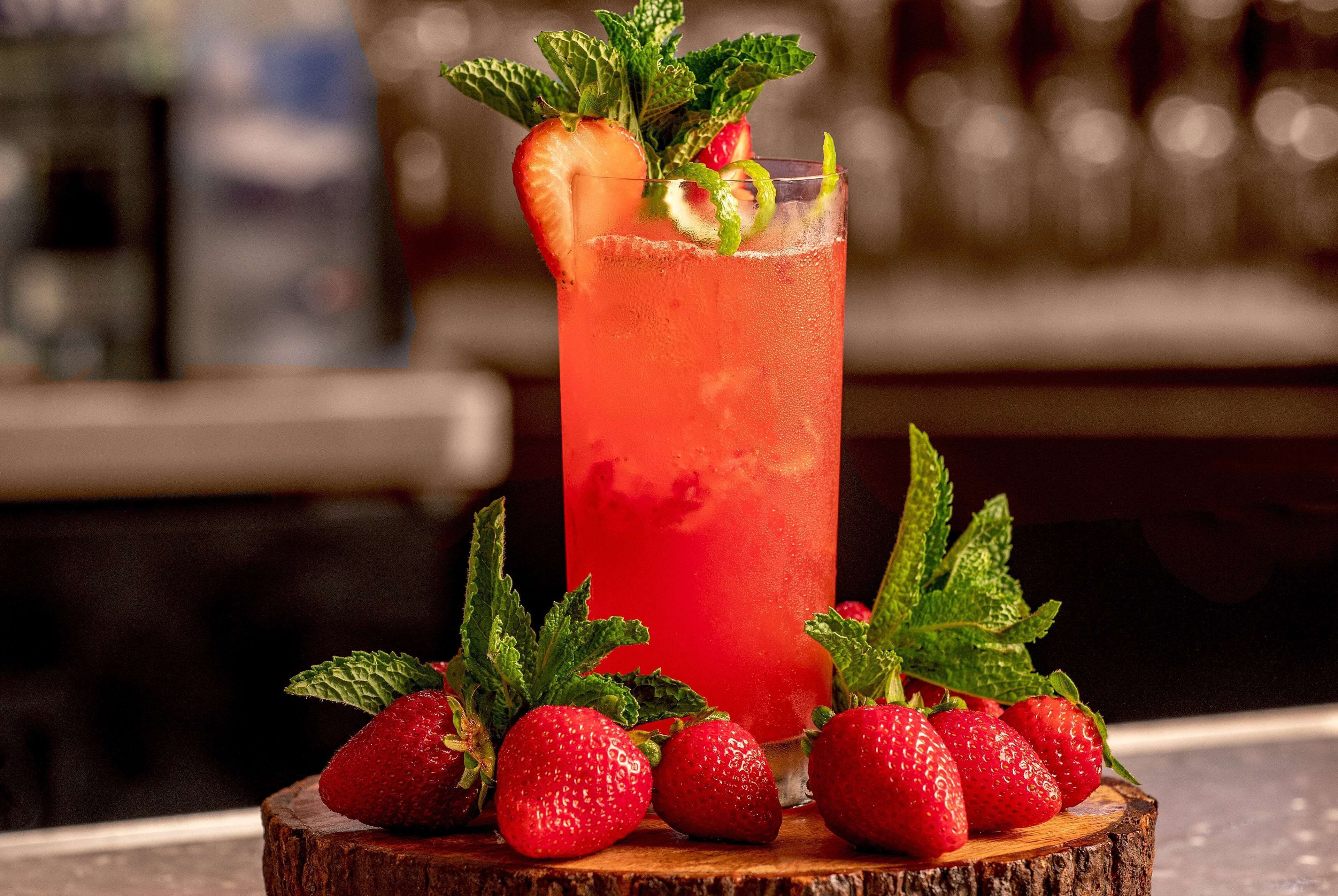 A tall glass of red beverage garnished with a strawberry and mint leaves, surrounded by fresh strawberries on a wooden surface.