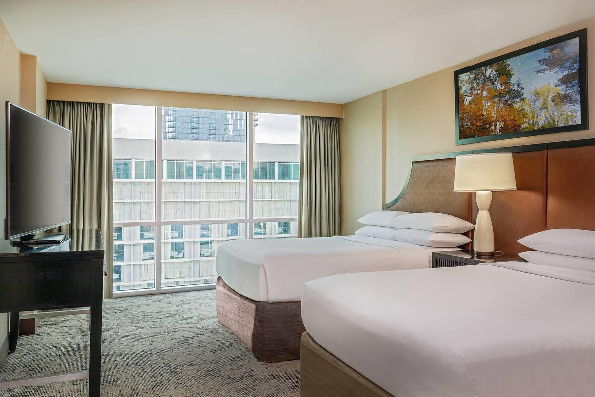 A hotel room with two beds, a TV, a desk, a lamp, and a large window with a city view.