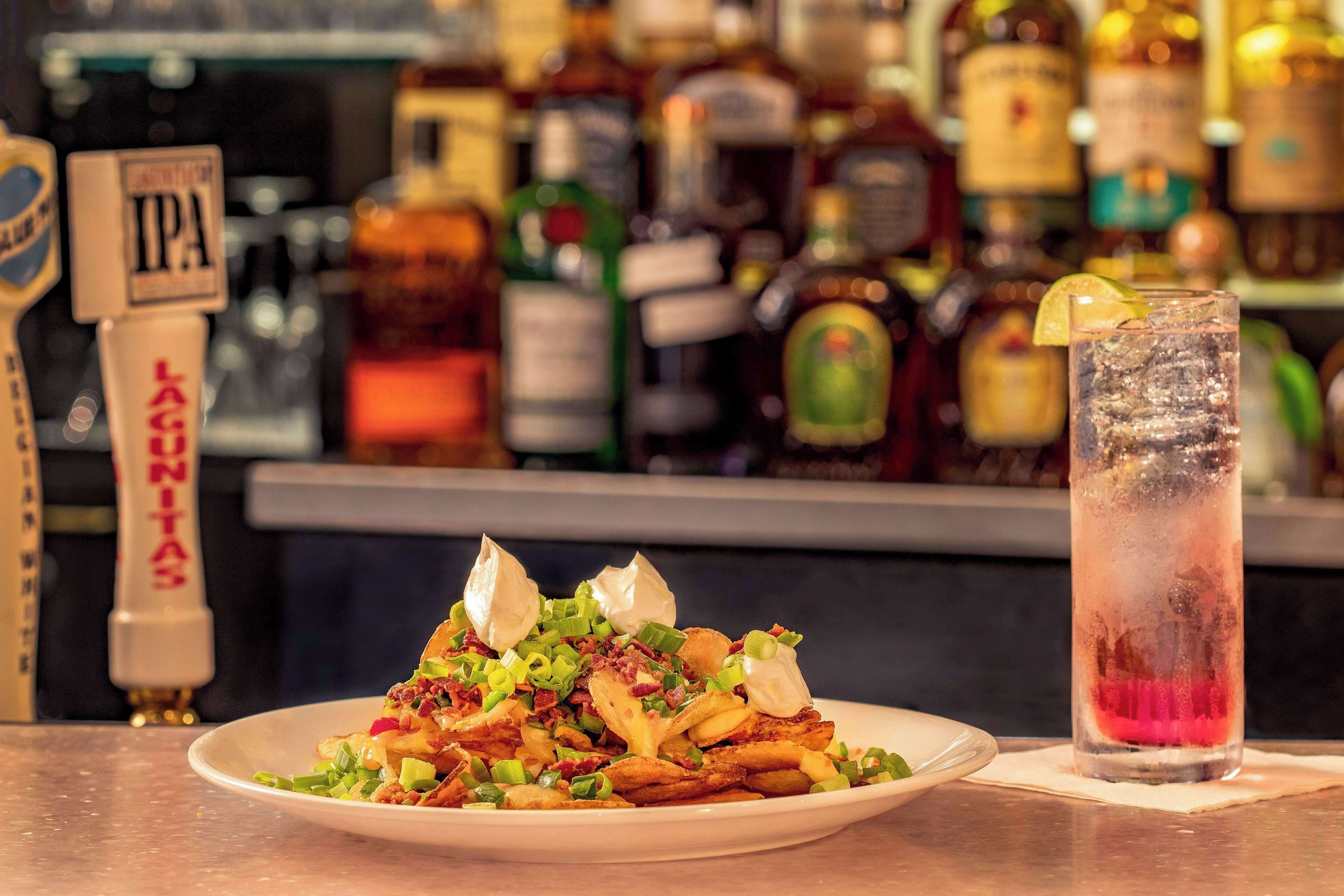 A plate of loaded fries with toppings and a glass of iced drink with a slice of lime on a bar counter.