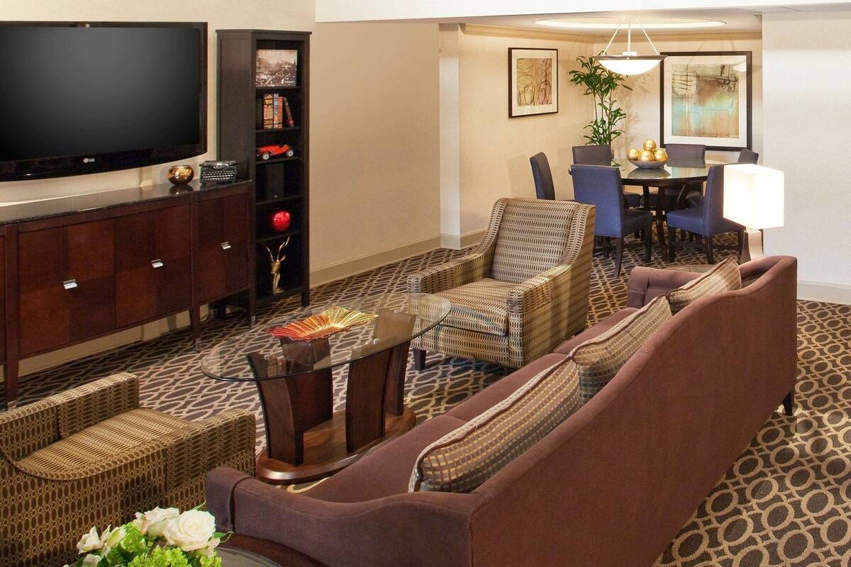 A hotel living room with a flat-screen TV, a bookshelf, a dining area with a table and chairs, and a patterned carpet.