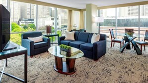 A modern living room with a sofa, armchairs, a coffee table, and a TV. Large windows offer a view of the cityscape. - Courtland Grand Hotel, Trademark Collection by Wyndham (Atlanta)