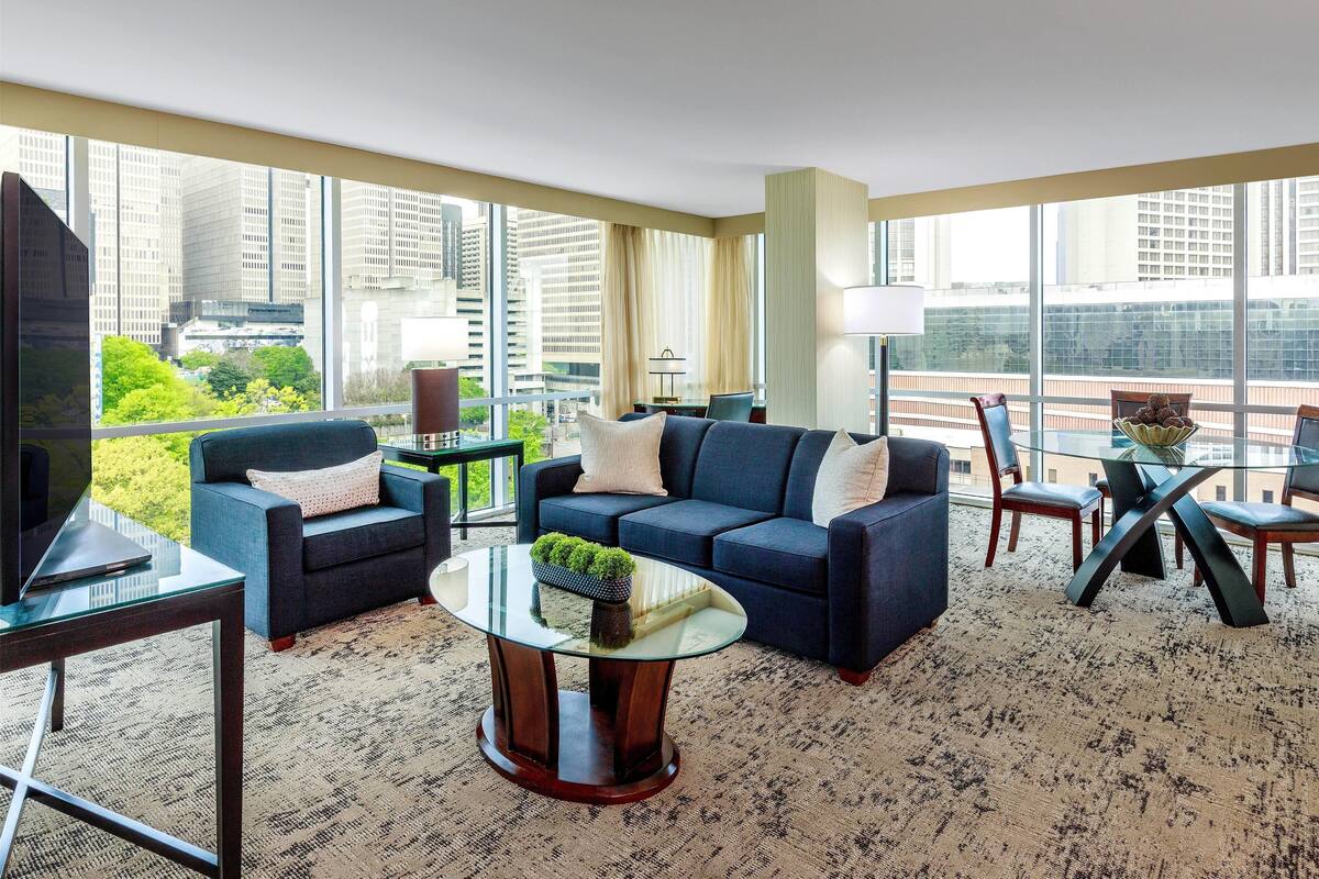 A modern living room with a sofa, armchairs, a coffee table, and a TV. Large windows offer a view of the cityscape.