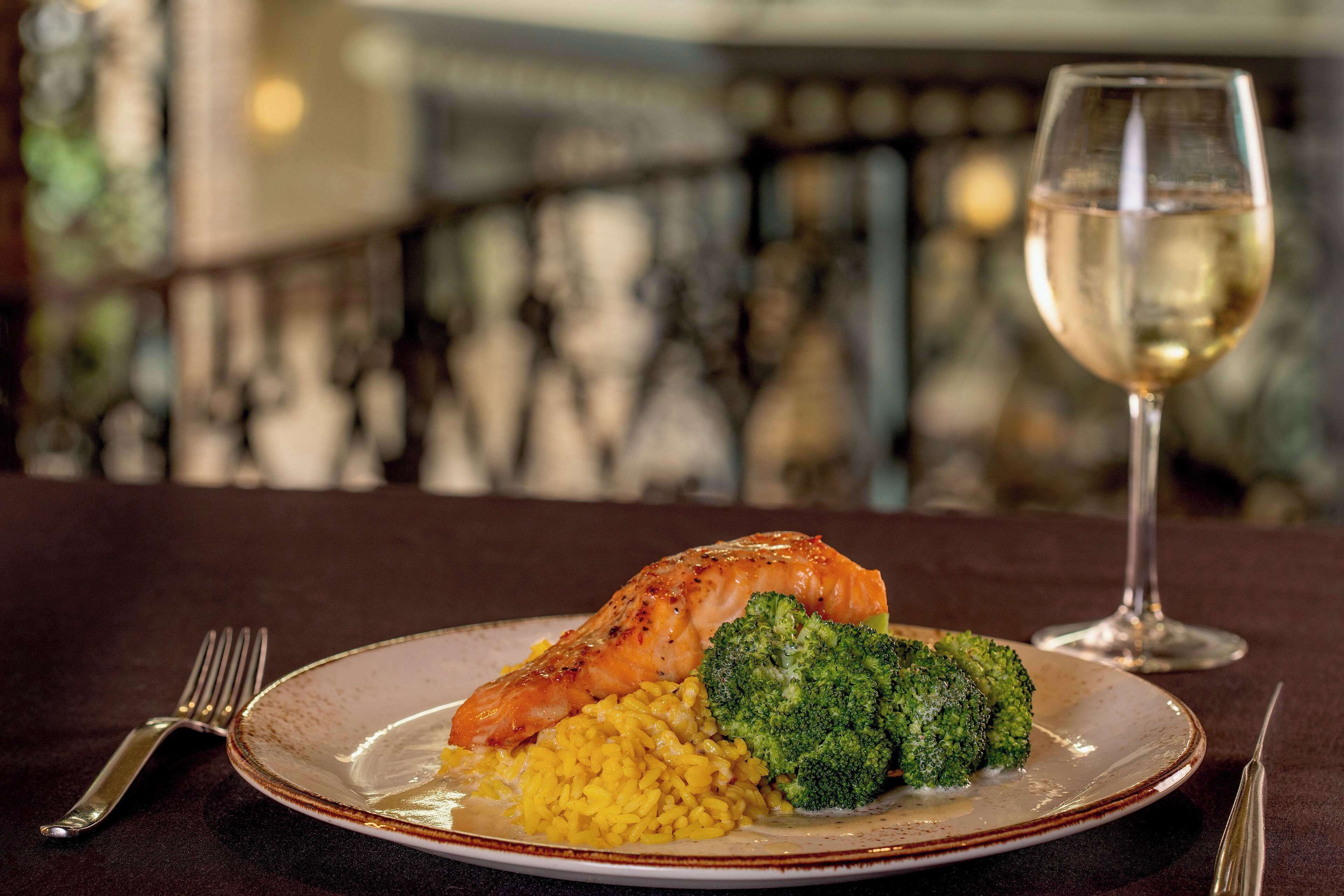 Een bord met zalm, broccoli en rijst, vergezeld van een glas witte wijn, op een donkere tafeldoek.