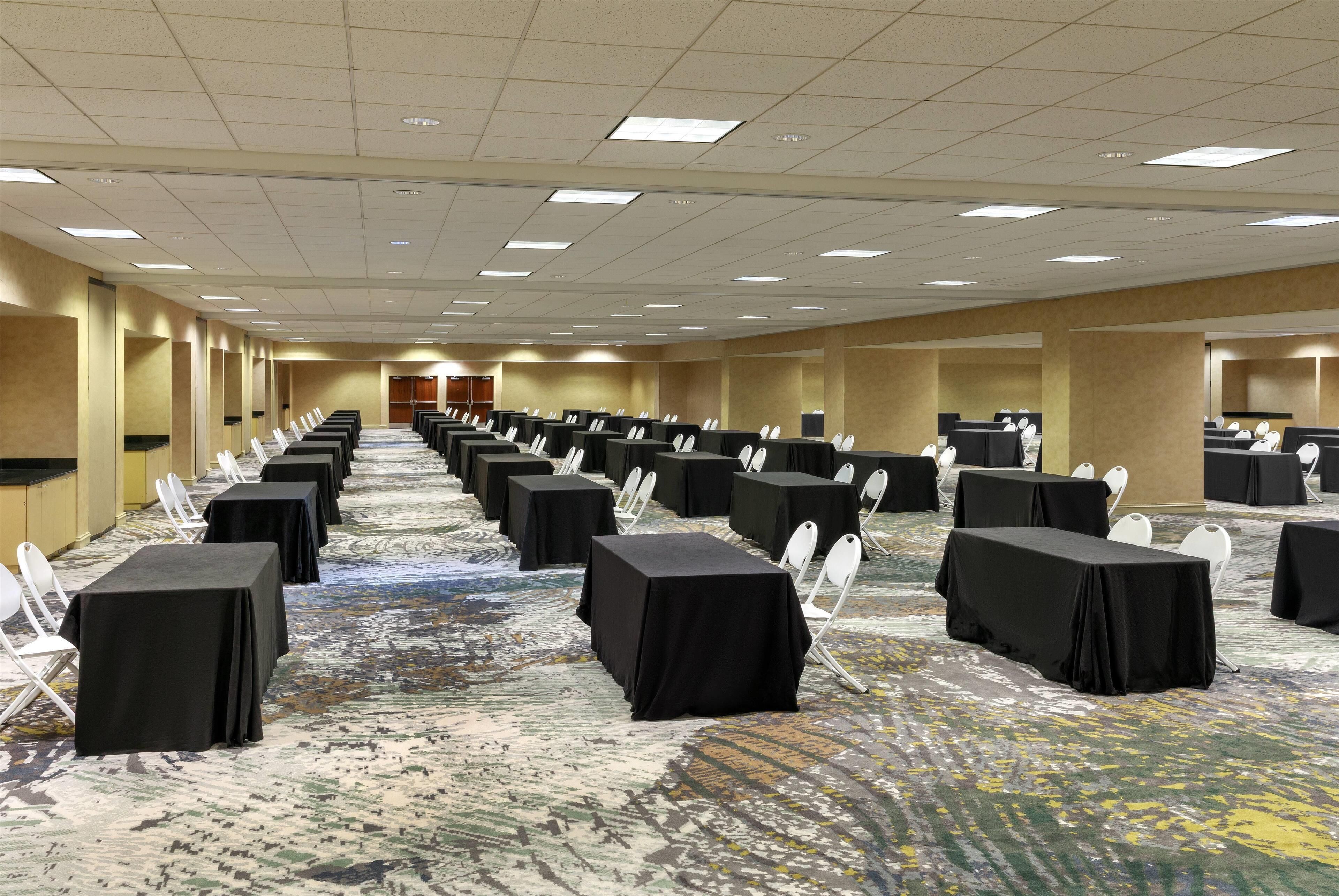 Een ruime congreszaal met rijen tafels en stoelen, een vloerbedekking en een hoog plafond met inbouwverlichting.
