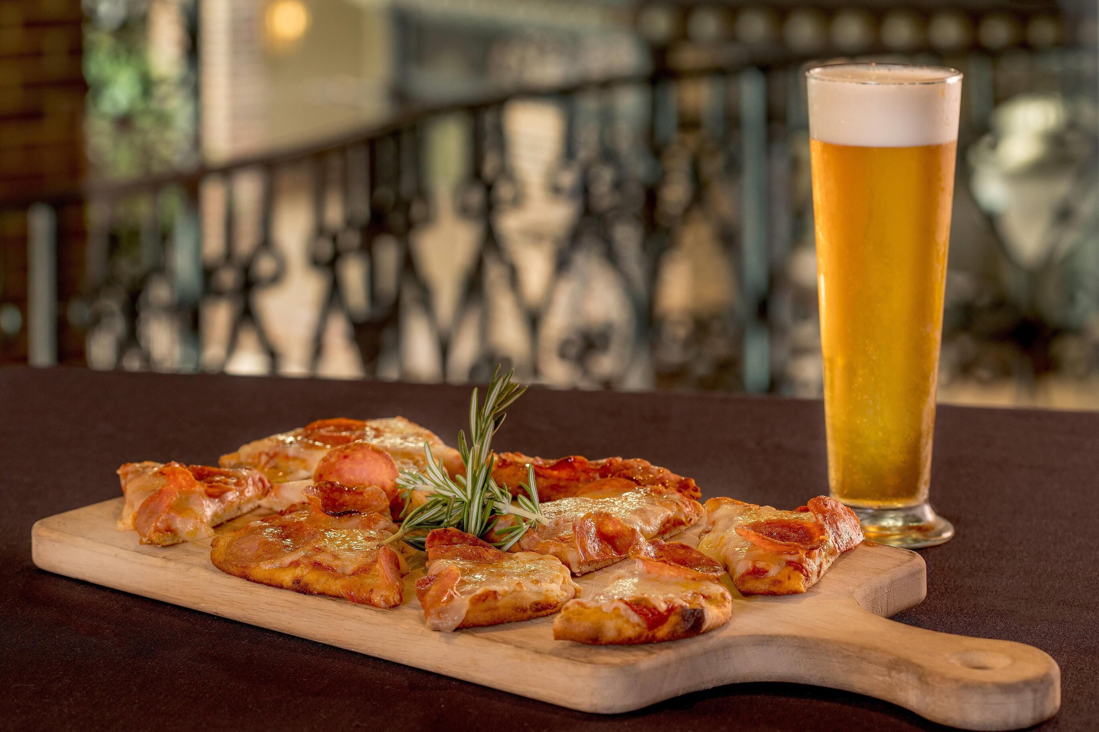 A wooden cutting board with pizza slices and a sprig of rosemary, accompanied by a tall glass of beer.