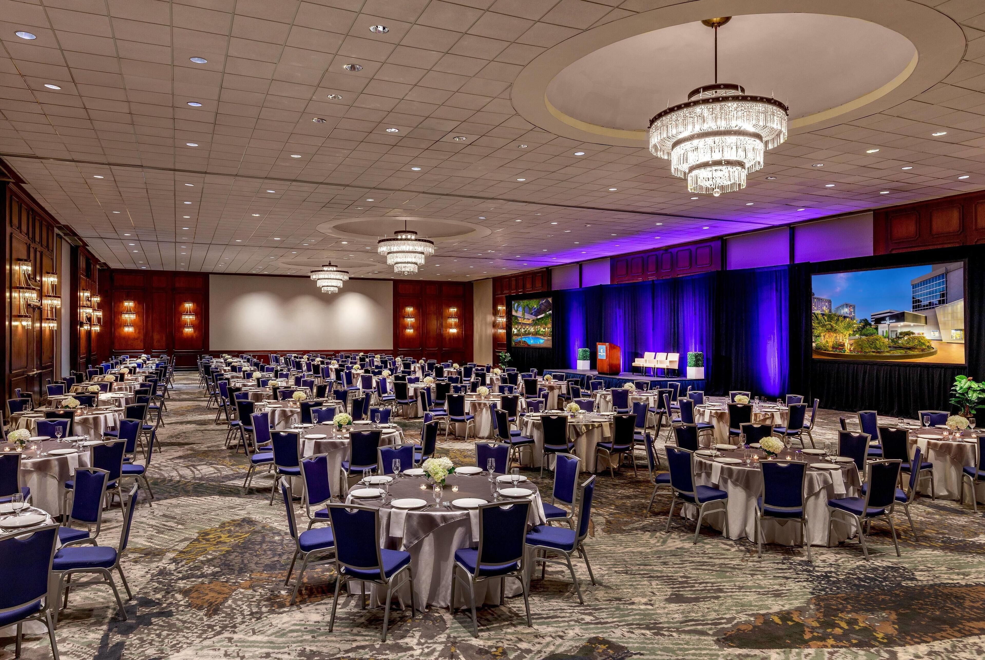 A large banquet hall with round tables and chairs, a stage with a large screen, and chandeliers.