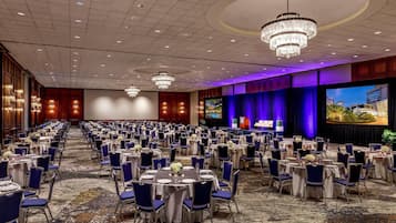 Un amplio salón de banquetes con mesas y sillas redondas, un escenario con una pantalla grande y candelabros.