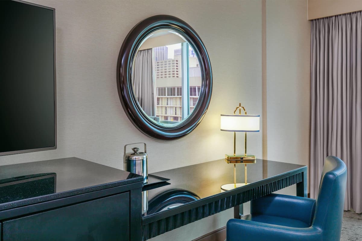 A hotel room with a large mirror, a desk with a lamp, a blue chair, and a view of buildings through the window.