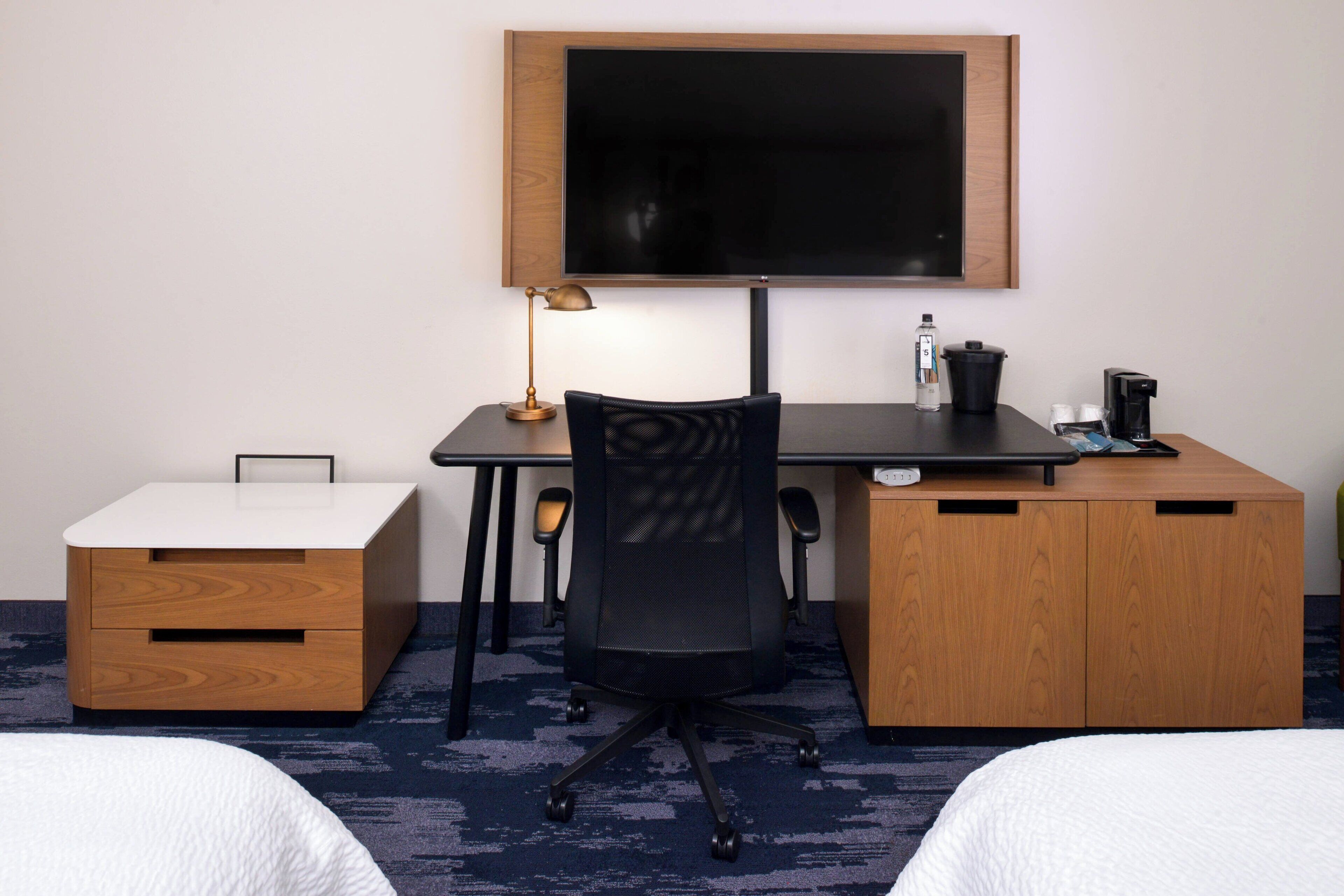 A hotel room with a desk, two beds, a TV, a lamp, a coffee maker, and a small cabinet.