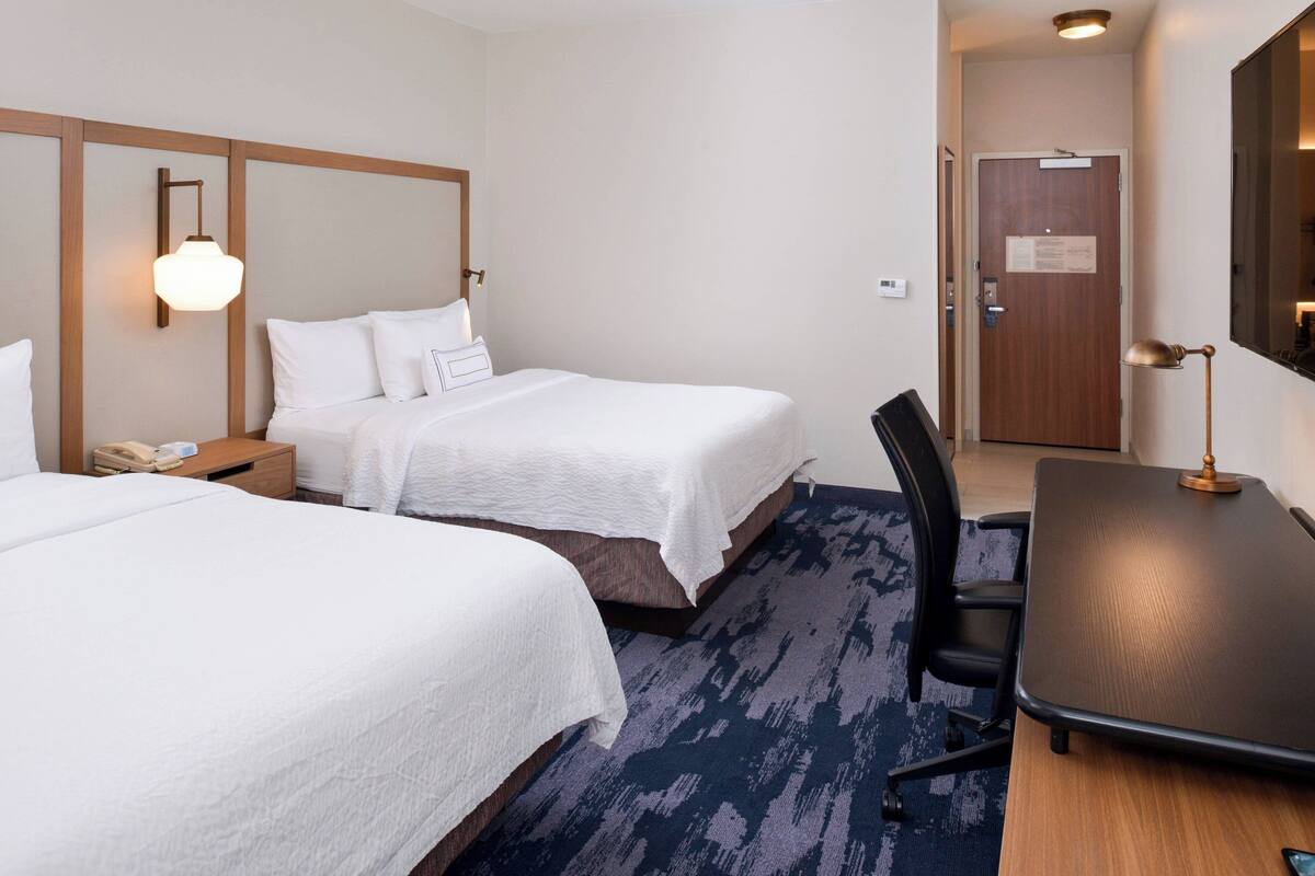 A hotel room with two beds, a desk, a chair, a lamp, and a door.