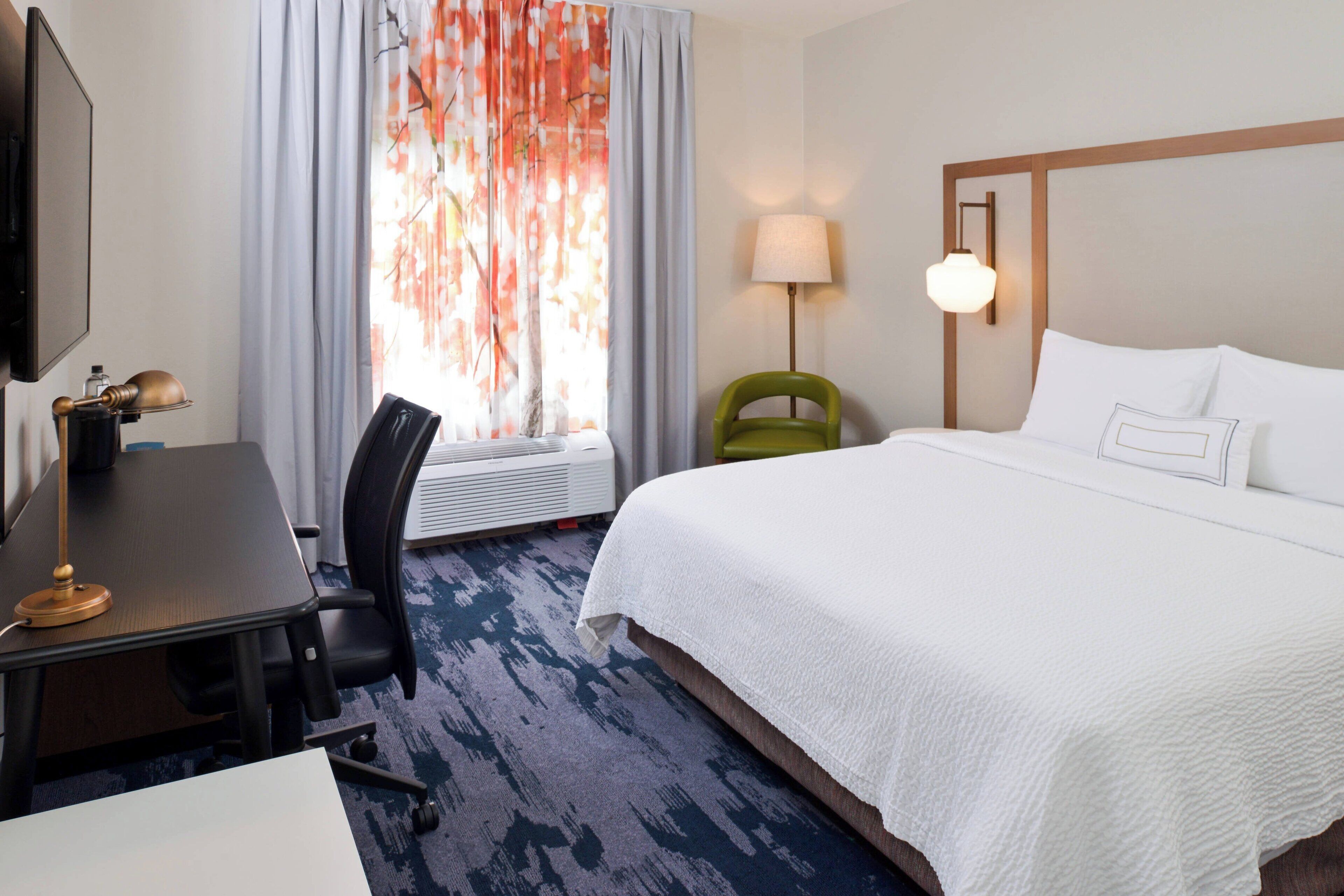 Une chambre d’hôtel avec un lit, un bureau, une chaise, une lampe et une fenêtre avec des rideaux.
