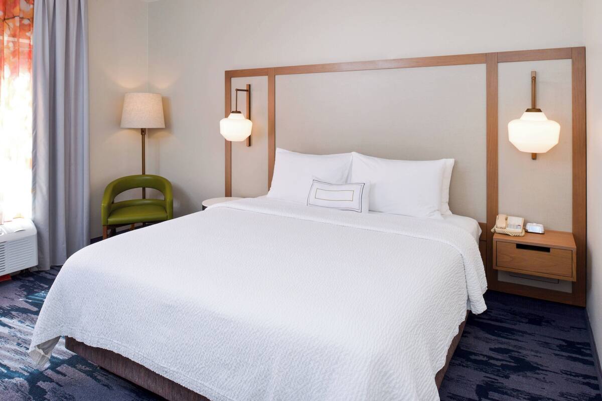 A neatly made bed with white linens, a green chair, a bedside table with a lamp, and a wall-mounted phone.
