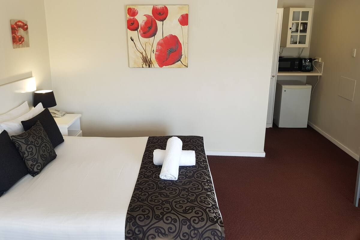 A hotel room with a bed, a bedside table with a lamp, a painting of red flowers on the wall, and a kitchenette in the background.
