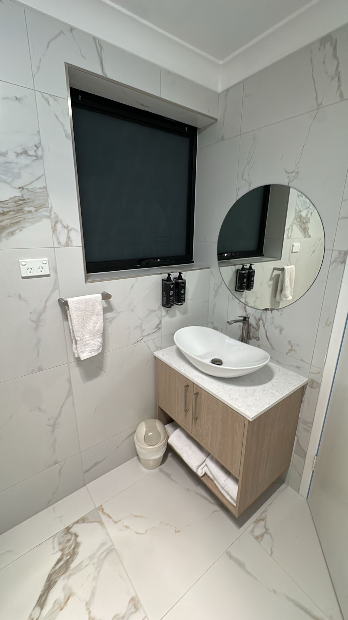 A bathroom with a white sink, wooden vanity, marble walls, a black-framed mirror, and a black TV mounted above the sink.