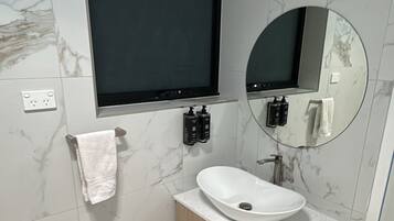 A bathroom with a white sink, wooden vanity, marble walls, a black-framed mirror, and a black TV mounted above the sink.