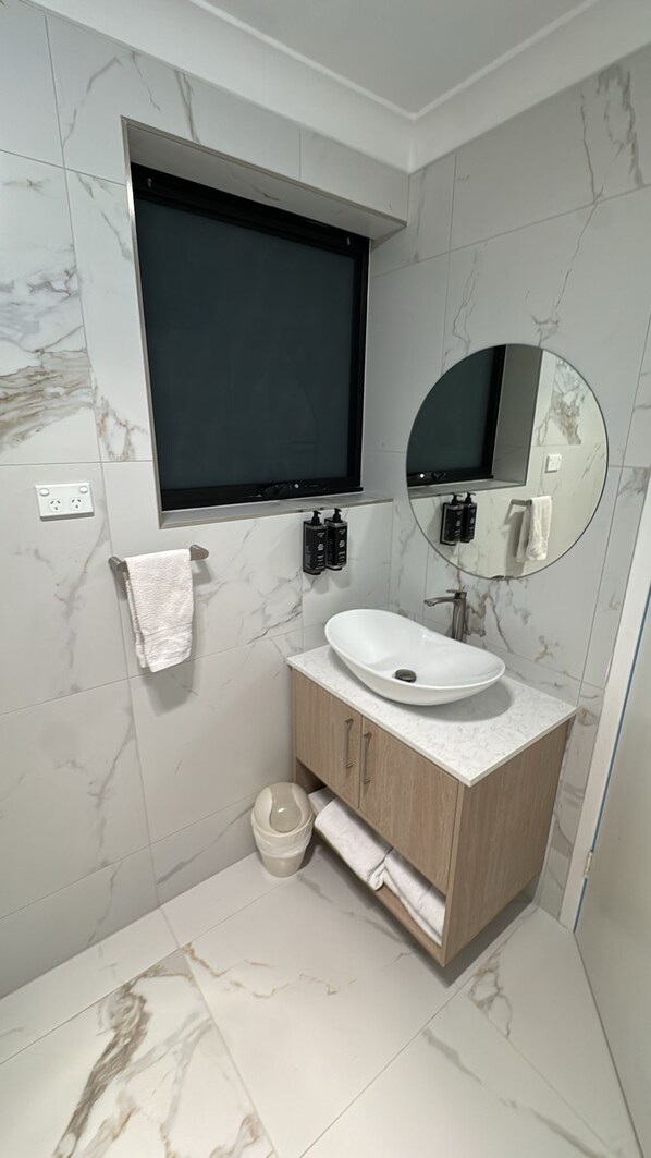 A bathroom with a white sink, wooden vanity, marble walls, a black-framed mirror, and a black TV mounted above the sink. - Clare Valley Motel (Clare)