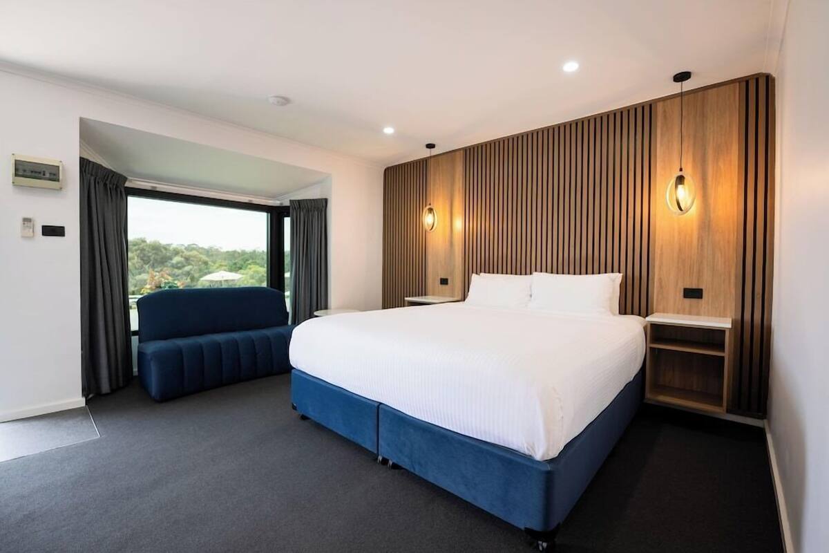 A modern hotel room with a large bed, a seating area, and a view through a window.