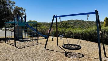 Un parque infantil con tobogán, columpios y un neumático colgante sobre suelo de grava.