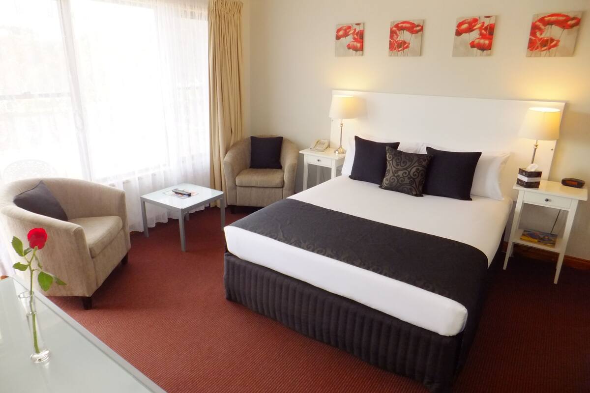 A hotel room with a large bed, a chair, a small table, a lamp, and artwork on the wall.